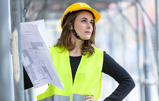 Ein Porträt einer Bauleiterin, die mit persönlicher Schutzausrüstung wie Schutzhelm und Warnweste auf einer Baustelle steht.