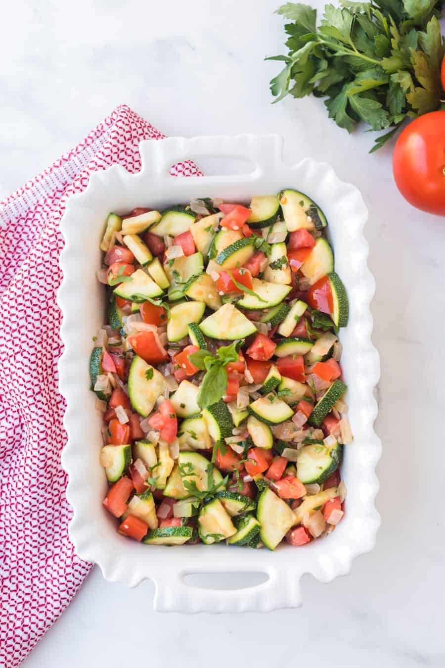 zucchini and tomato side dish in a white dish with pink and white towel and red tomato in background.