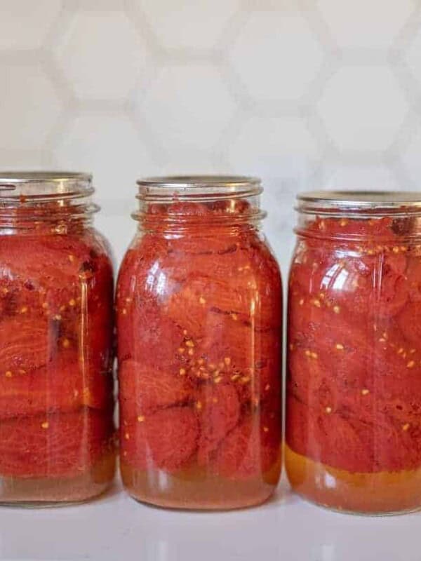 Three glass jars filled with canned tomatoes, preserved using the water bath method, are lined up on a white countertop. The jars have silver lids, and the background features a hexagonal tile pattern.