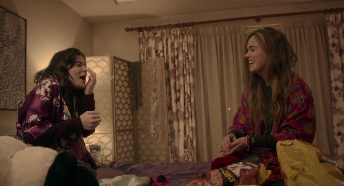 Two best friends laughing together, sitting on a bed with snacks in a cozy room.