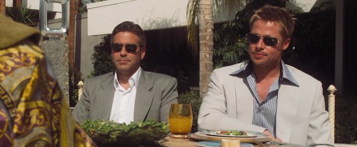 Two men in suits and sunglasses sitting at an outdoor table, embodying an iconic best friend duo.