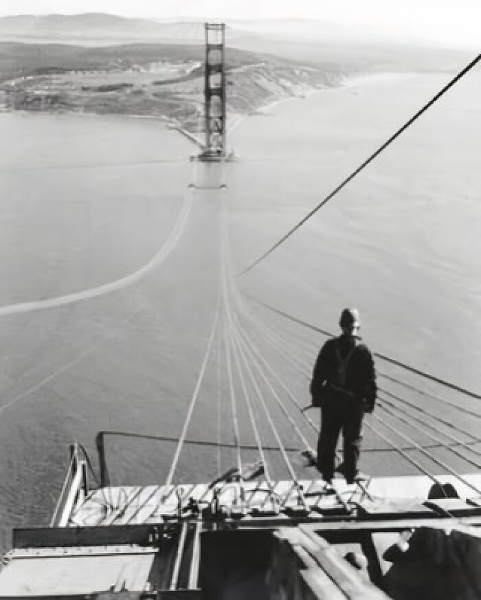 Worker standing on the Golden Gate Bridge under construction, with cables stretching across the bay, highlighting historical architecture.