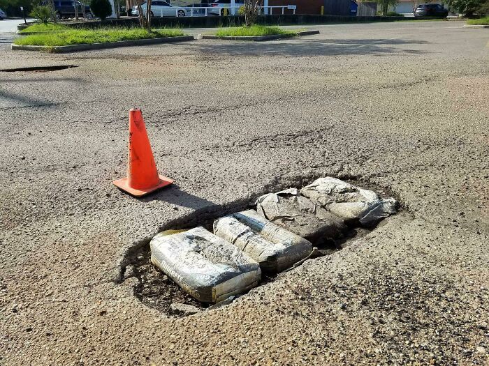 Traffic cone beside a pothole filled with cement bags, highlighting lazy-infuriating-people approach to road repair.