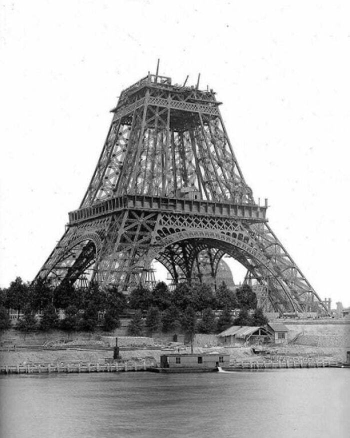 Eiffel Tower under construction, showcasing historical progress amidst a backdrop of trees and water.