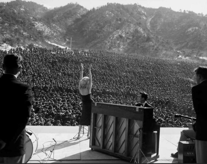 Performer on stage in front of a large crowd, historical setting with mountains in the background.