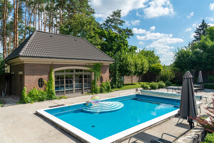 Backyard pool with lounge chairs and a modern guest house surrounded by trees in the neighbors' yard.