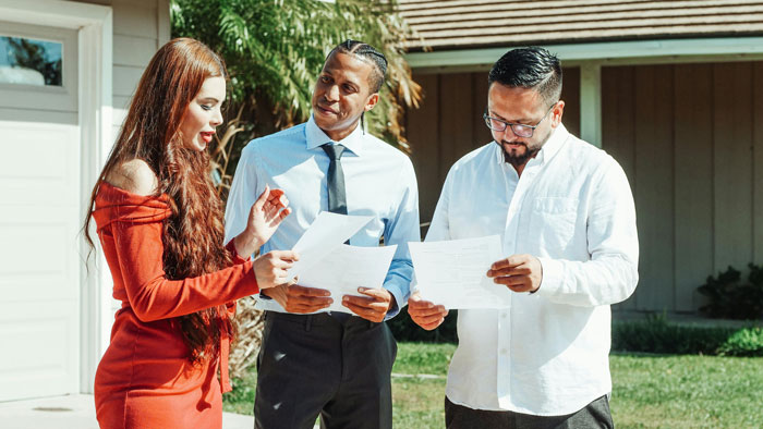 Three people discussing documents in a neighbor's yard, with two men wearing shirts and a woman in a red dress.