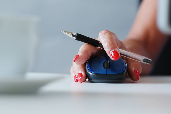 A hand with red nails, holding a pen and a blue computer mouse, suggesting multitasking.