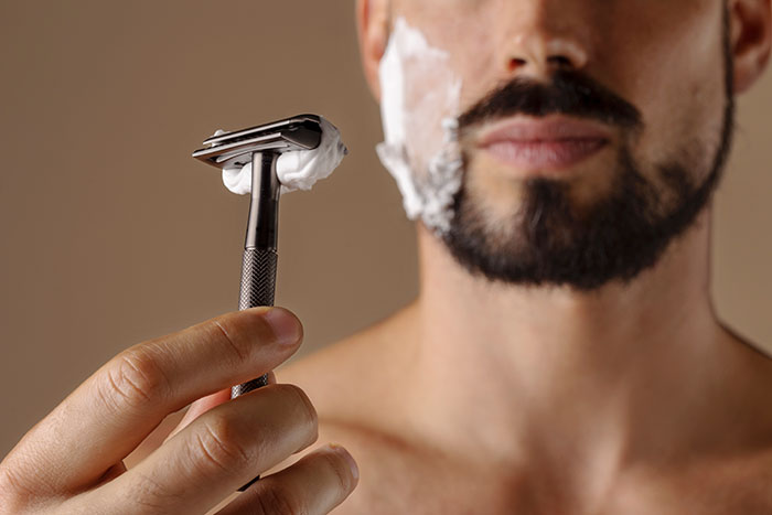 Man holding a razor with shaving cream on his face, focusing on grooming technique.