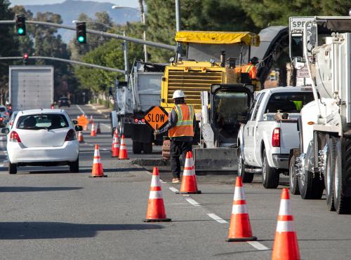 Wide view of road construction site and a man holding traffic sign that says SLOW .Bright orange traffic cones and trucks and cars are visible