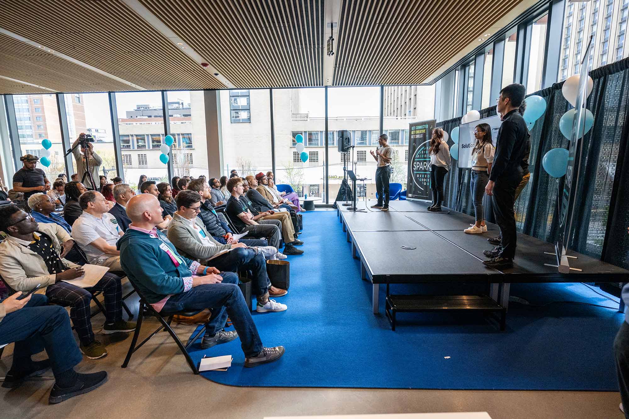 Photo: Students present their apps to judges, stakeholders, and visitors May 2 at CDS for BU Spark! Demo Day. They sit in a large room with floor to ceiling windows.