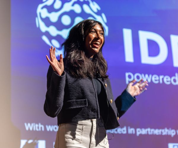 A woman gestures excitedly while presenting in front of a colorful backdrop at a large event