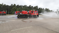 Ein rotes Feuerwehrfahrzeug fährt über eine Betonfläche und sprüht dabei Wasser