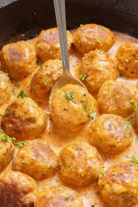 A skillet filled with cooked meatballs in a creamy sauce, garnished with chopped herbs, with a serving spoon resting among the meatballs.