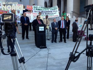 A group of people stand on the steps of a building, some holding signs advocating for the decriminalization of cannabis. Cameras on tripods are positioned in the foreground, capturing the scene. A speaker addresses the gathering from a podium. CA Norml