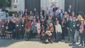 A diverse group of people, including individuals with mobility aids, stands smiling on steps in front of a grand building. Many wear lanyards, hinting at a Cal NORML conference or event gathering. The backdrop features large, decorative doors celebrating their accomplishments. CA Norml