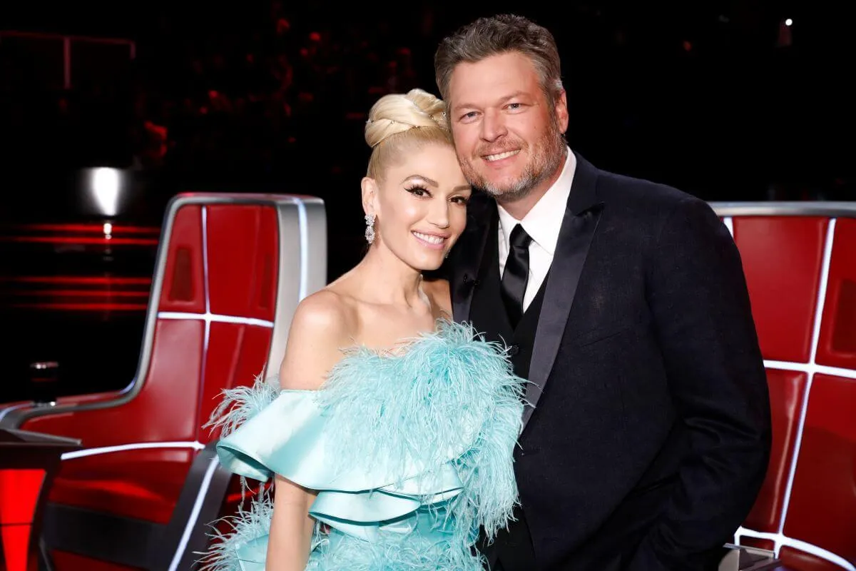 Gwen Stefani wears a blue dress and stands with Blake Shelton, who wears a suit. They stand in front of two red chairs.