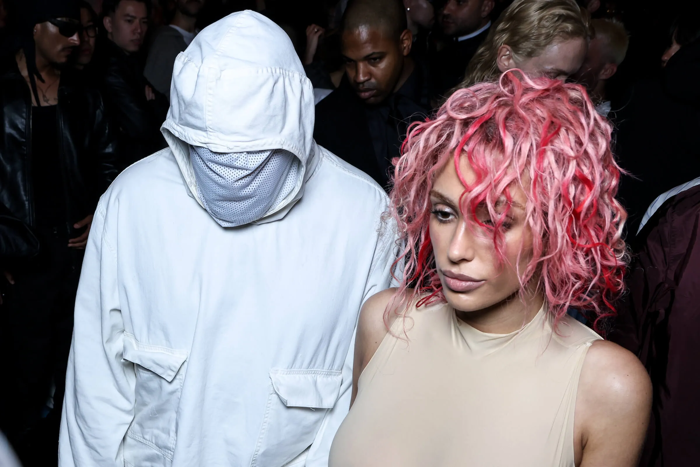 Kanye West and Bianca Censori at a fashion show. West is in a head-to-toe white outfit that covers his face. Censori is wearing a nude bodysuit with pink hair.