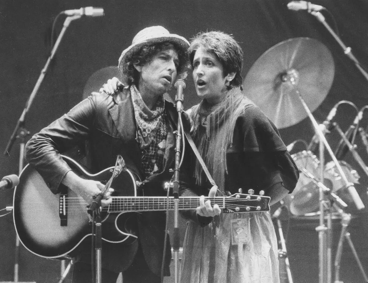 A black and white picture of Bob Dylan and Joan Baez singing into the same microphone. He plays a guitar.