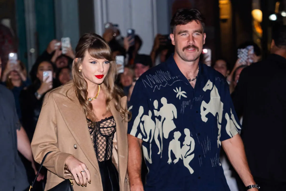 Taylor Swift and Travis Kelce walking hand in hand in New York City at night. Swift wears a tan coat and Kelce wears a short-sleeve button-down shirt.