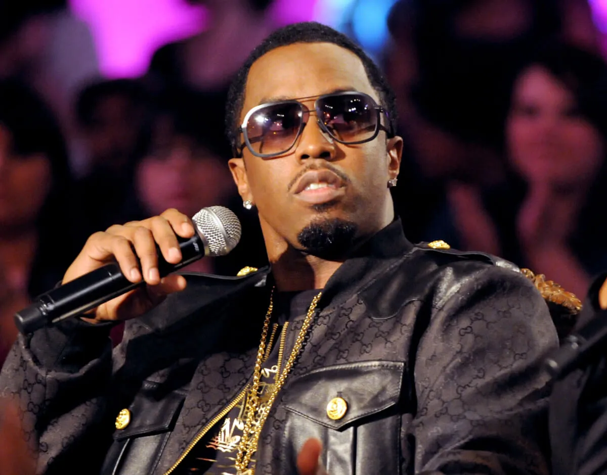 Sean 'Diddy' Combs wears a black jacket and sunglasses on 'Making the Band.' He holds a microphone.