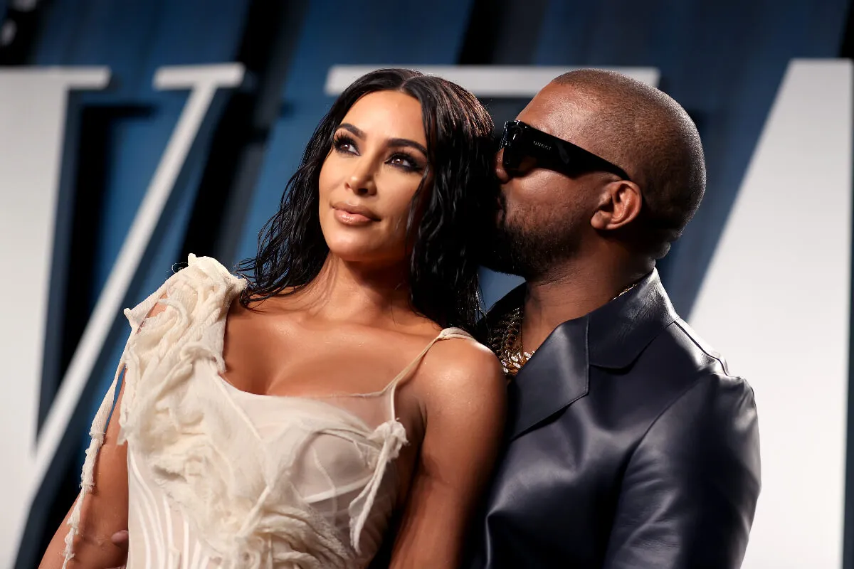 Kim Kardashian and Kanye West stand together. She wears a white dress and he wears a leather jacket and sunglasses.