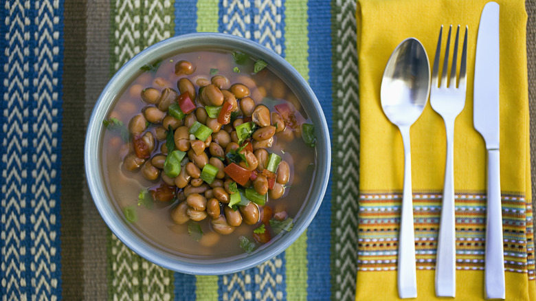 a bowl of beans sits on a colorful tablecloth next to silverware