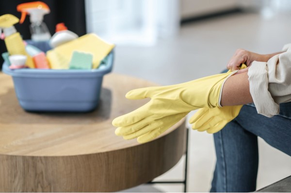 Person putting on yellow cleaning gloves with a basket of cleaning supplies in the background.