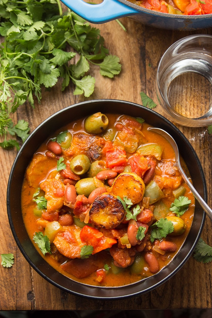 Wooden Table Set with a Bowl of Plantain Stew, Drinking Glass, Blue Pot, and Bunch of Cilantro