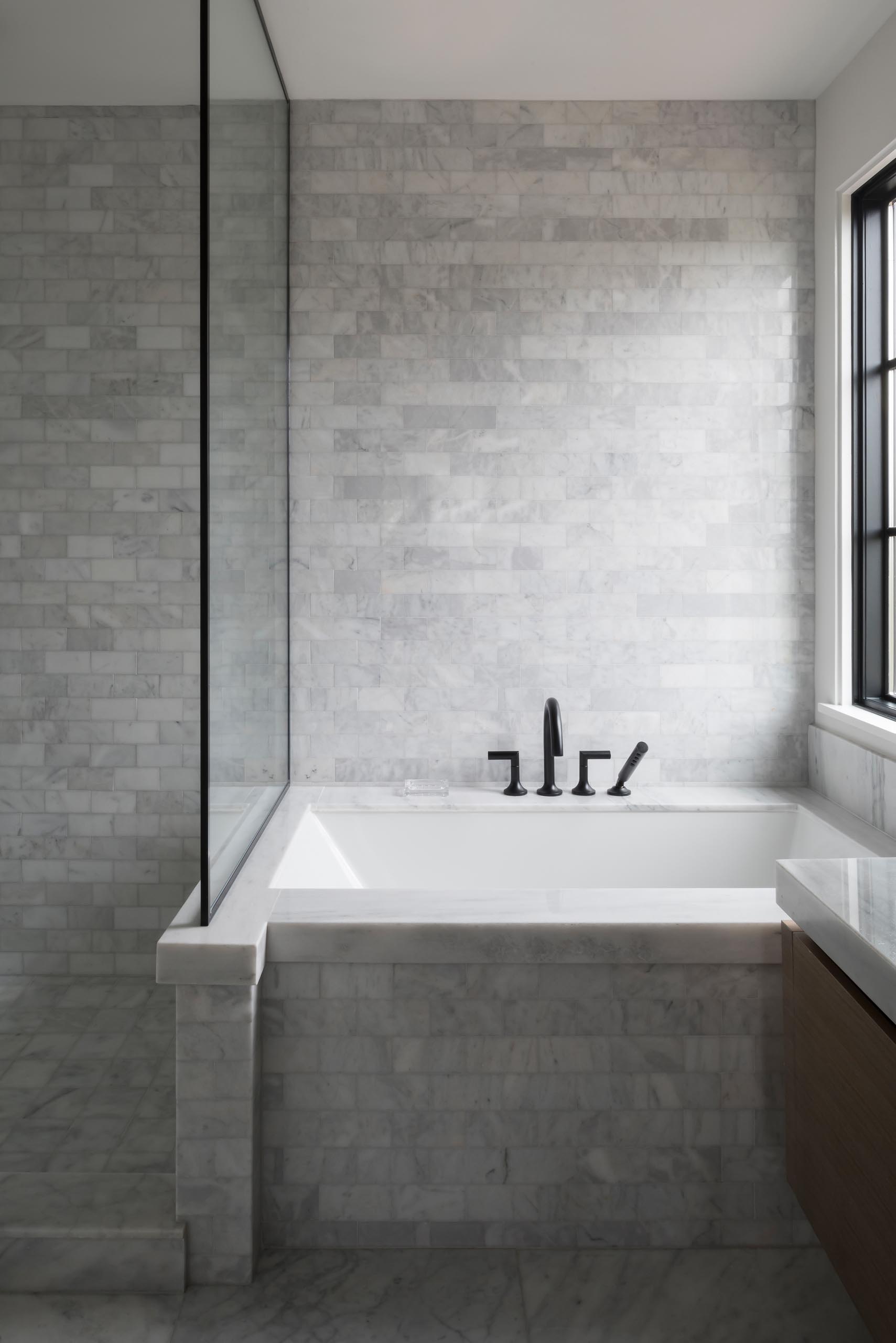 A modern bathroom with marble tiles, a built-in bathtub, and black framed glass shower screen.