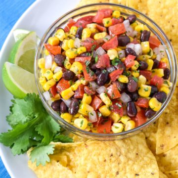 The black bean and corn salsa dip served in a glass bowl with corn tortilla chips, lime wedges and a cilantro sprig on a white plate.