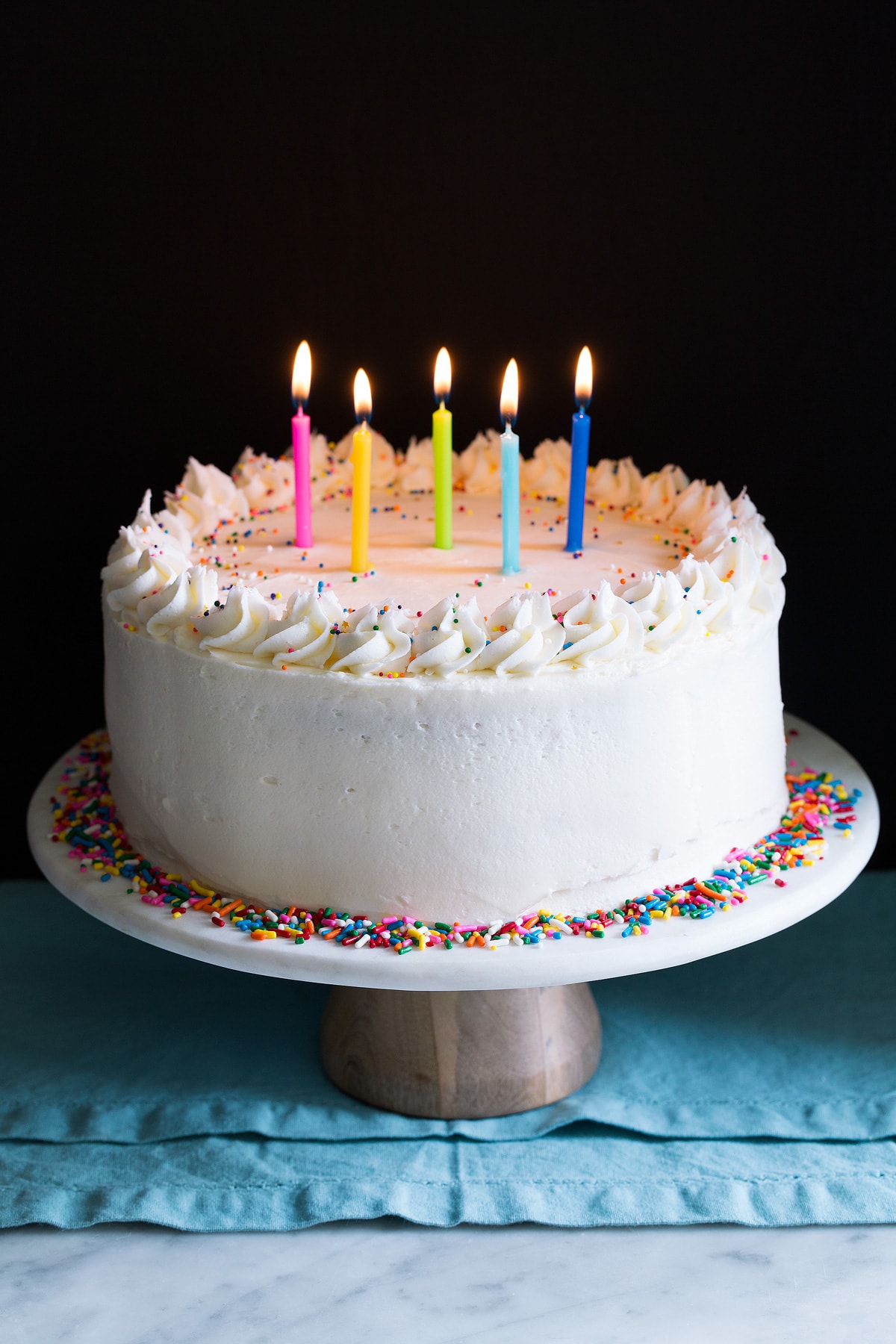 Whole birthday cake on a cake stand garnished with sprinkles and topped with lit birthday candles. 