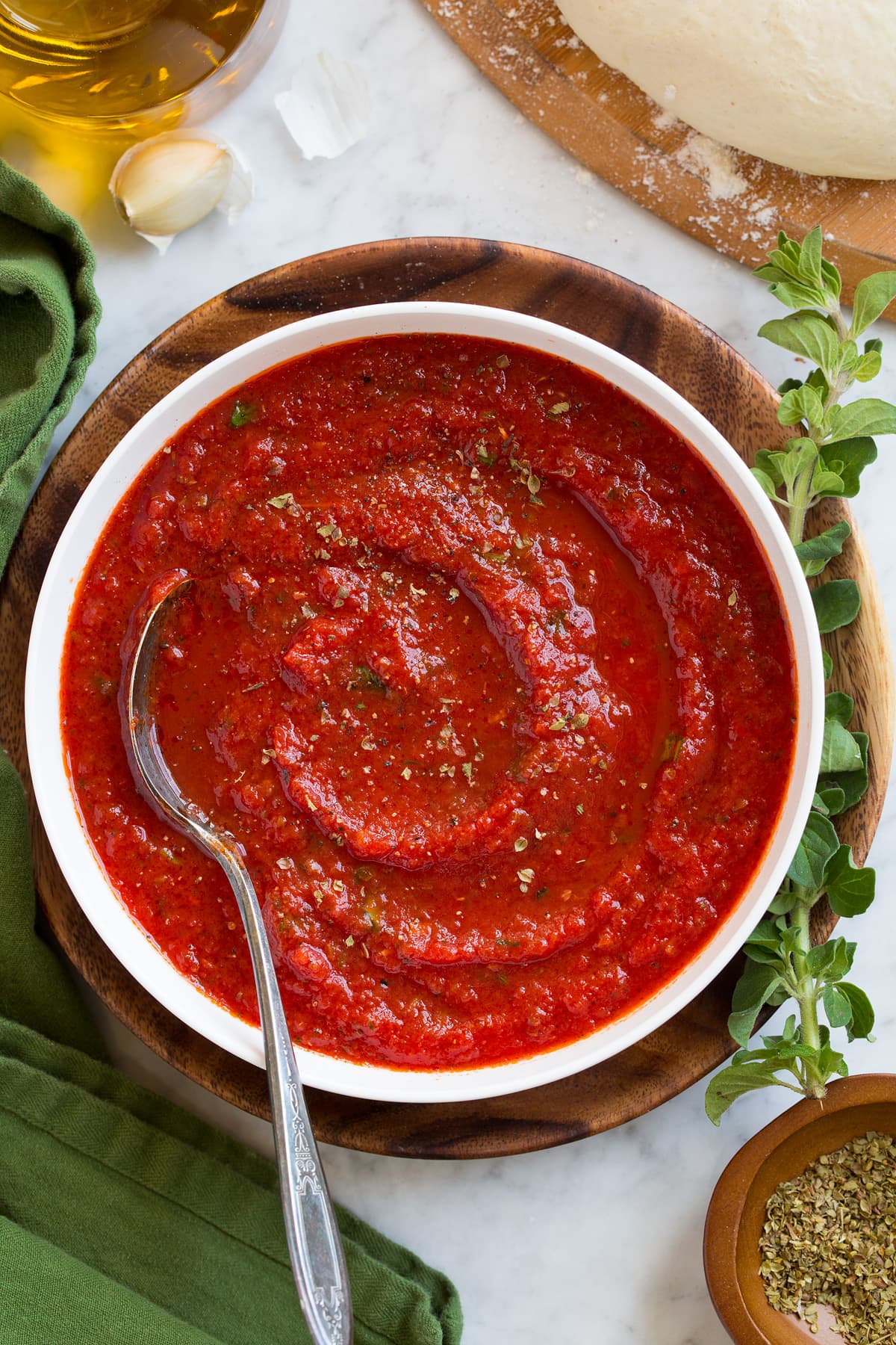 Overhead image of pizza sauce in a white bowl with a spoon in the sauce. Sauce is resting on a wooden plate over a marble surface.