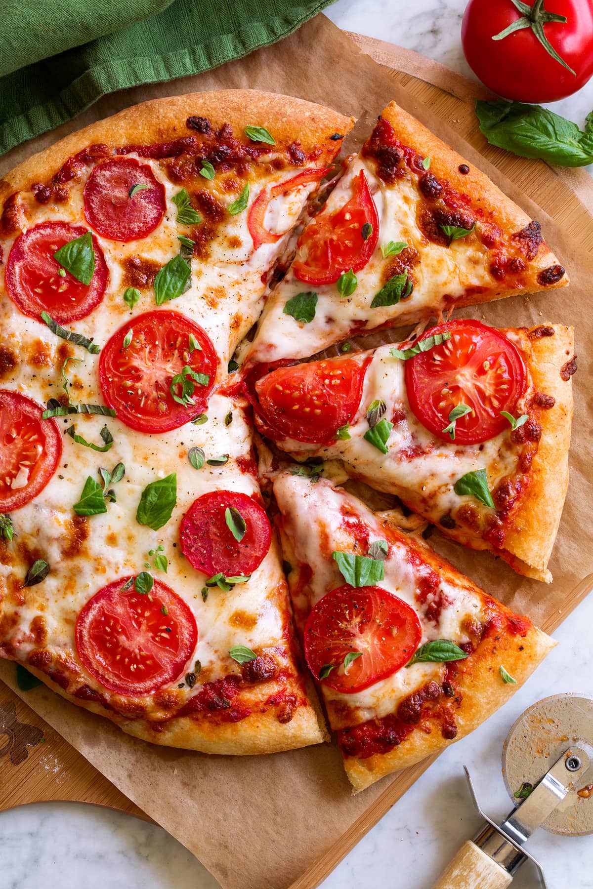 Pizza topped with red pizza sauce, mozzrella, tomatoes and herbs. It is cut into slices and sitting on parchment paper over a wooden pizza peel.