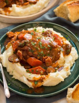 Coq au vin served over mashed potatoes on a green plate with bread to the side.