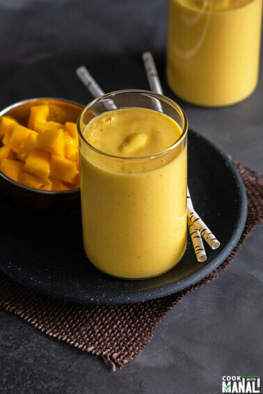 mango lassi served in a glass with 2 straws and another glass of mango lassi in the background