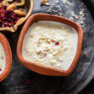 Rice Kheer served in a clay bowl and garnished with rose petals and a golden cast iron pan with nuts and rose petals in the background