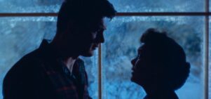 A man and a woman stood in front of a window at night look into each others' eyes  