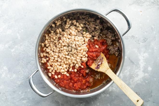 Sausage and bean stew ingredients being added together in a silver pot.