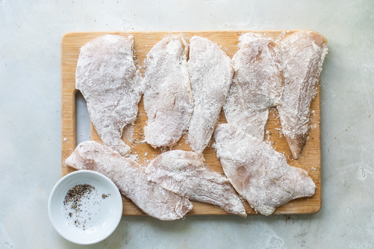 Chicken scallopini coated in flour on a wooden cutting board.