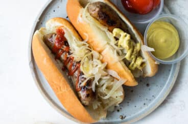 A plate of beer brats with ketchup, mustard, onions, and sauerkraut.