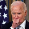 Joe Biden in a black suit puts his hand in front of his mouth