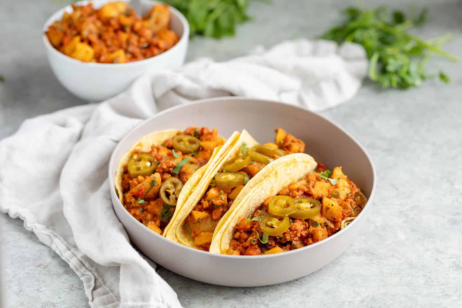 Three Tacos Filled With Vegan Chorizo And Diced Vegetables, Topped With Jalapeño Slices And Served In A Gray Bowl On A Light Gray Surface With A White Cloth And Parsley Nearby.