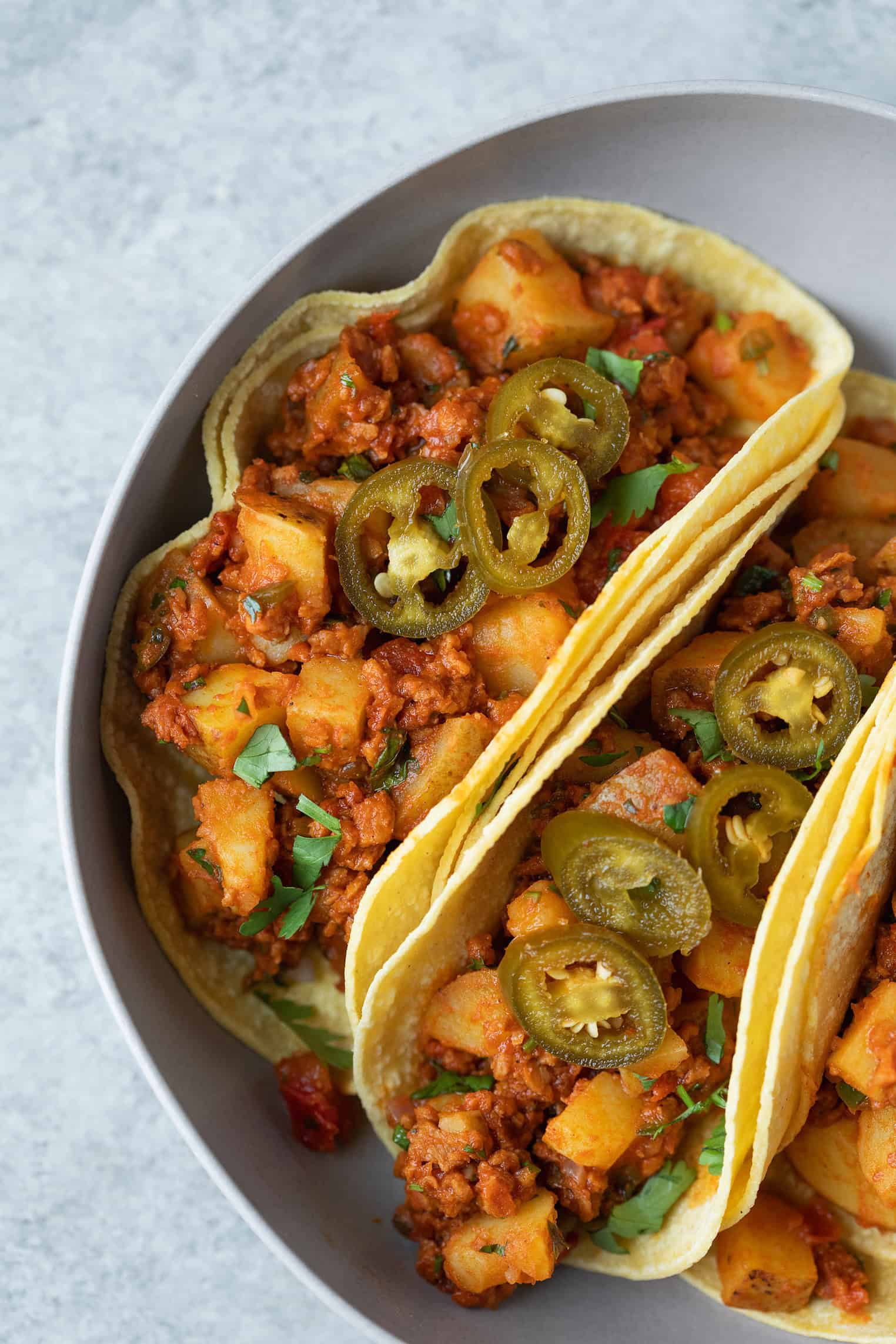 A Close-Up Image Of A Taco Filled With Spicy Potato And Chorizo Mixture, Garnished With Slices Of Jalapeño Peppers And Chopped Herbs, Served In A Gray Bowl.