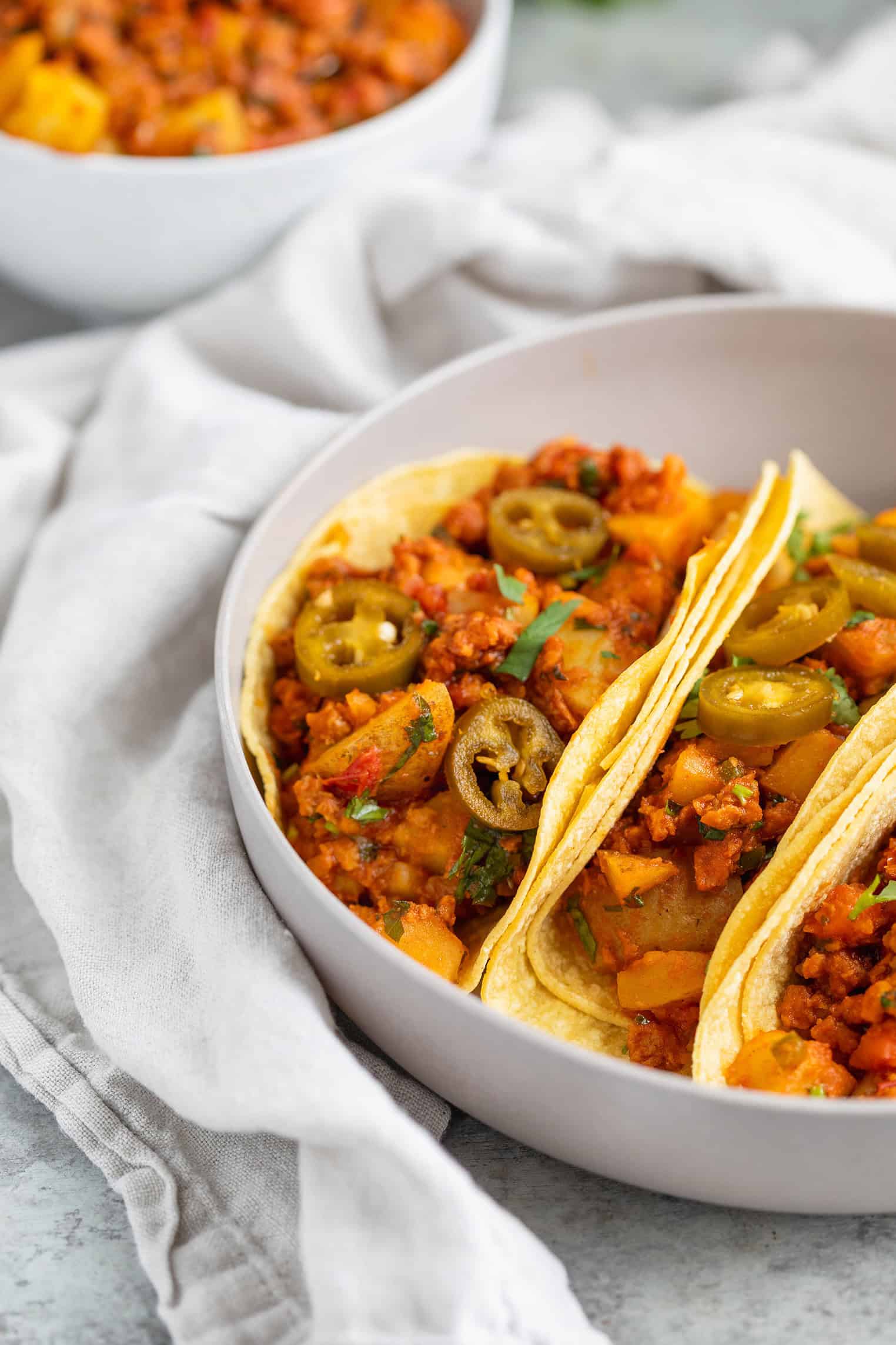 Three Soft Tacos Filled With Vegan Chorizo And Potatoes, Diced Vegetables, And Sliced Jalapeños, Garnished With Cilantro, Served In A White Bowl On A Marble Surface With A Cloth.