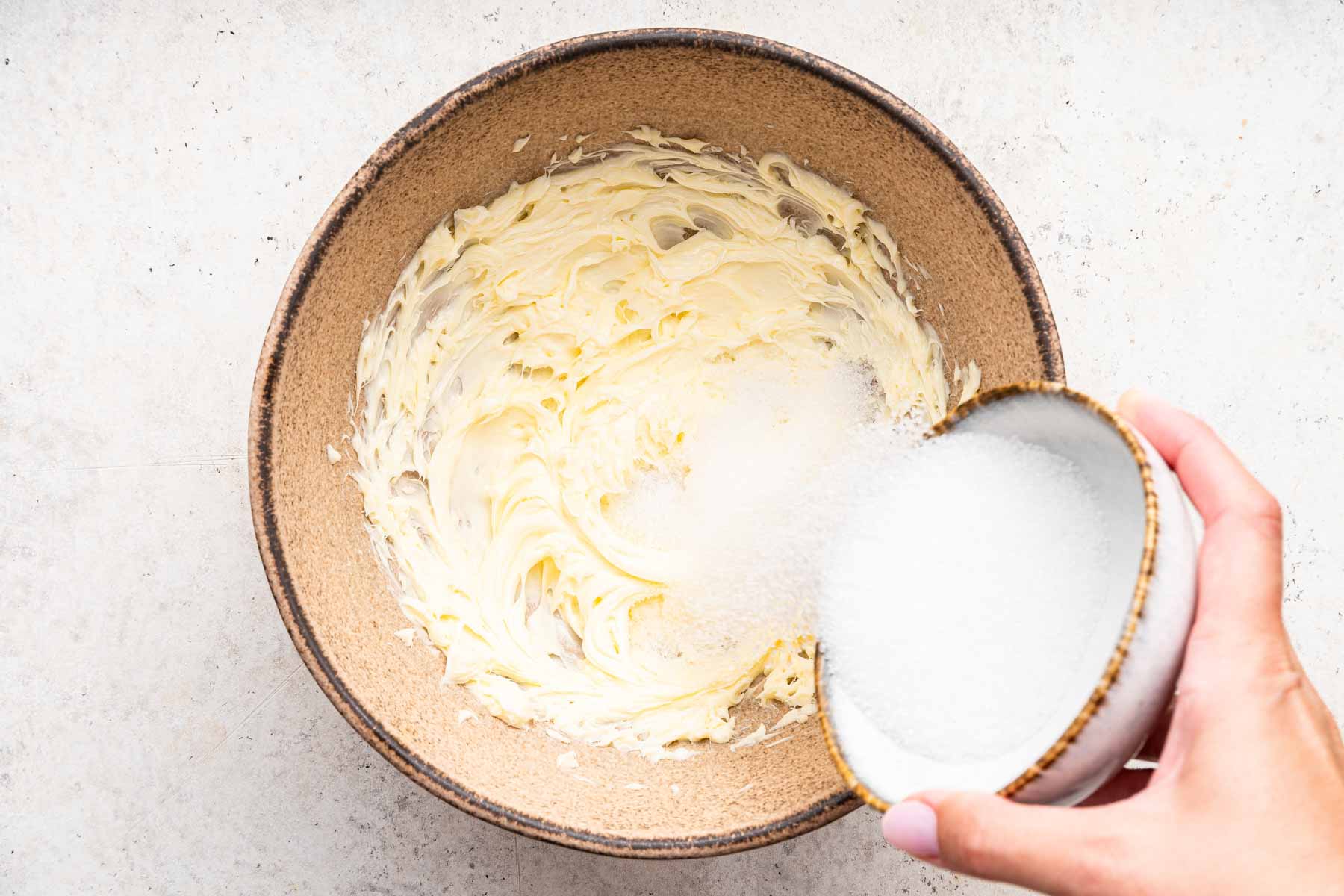 Hand pouring sugar into a brown bowl with butter.