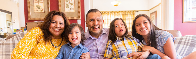 Young Latinx Family sitting on couch at home. One little boy has Down’s Syndrome.