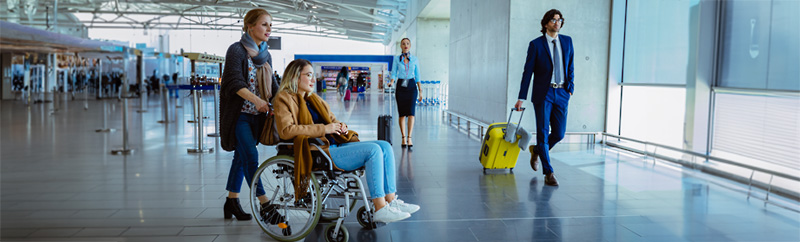 A women pushing a female friend with a disability in a wheel chair through the airport.