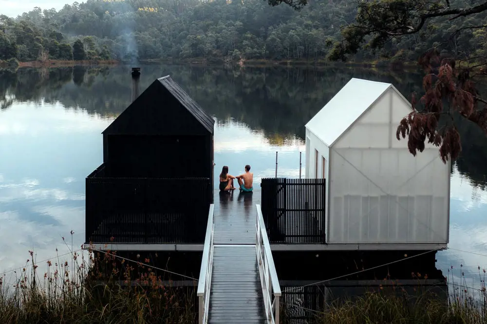 Bathers sitting on jetty between black and white sauna huts, overlooking lake.