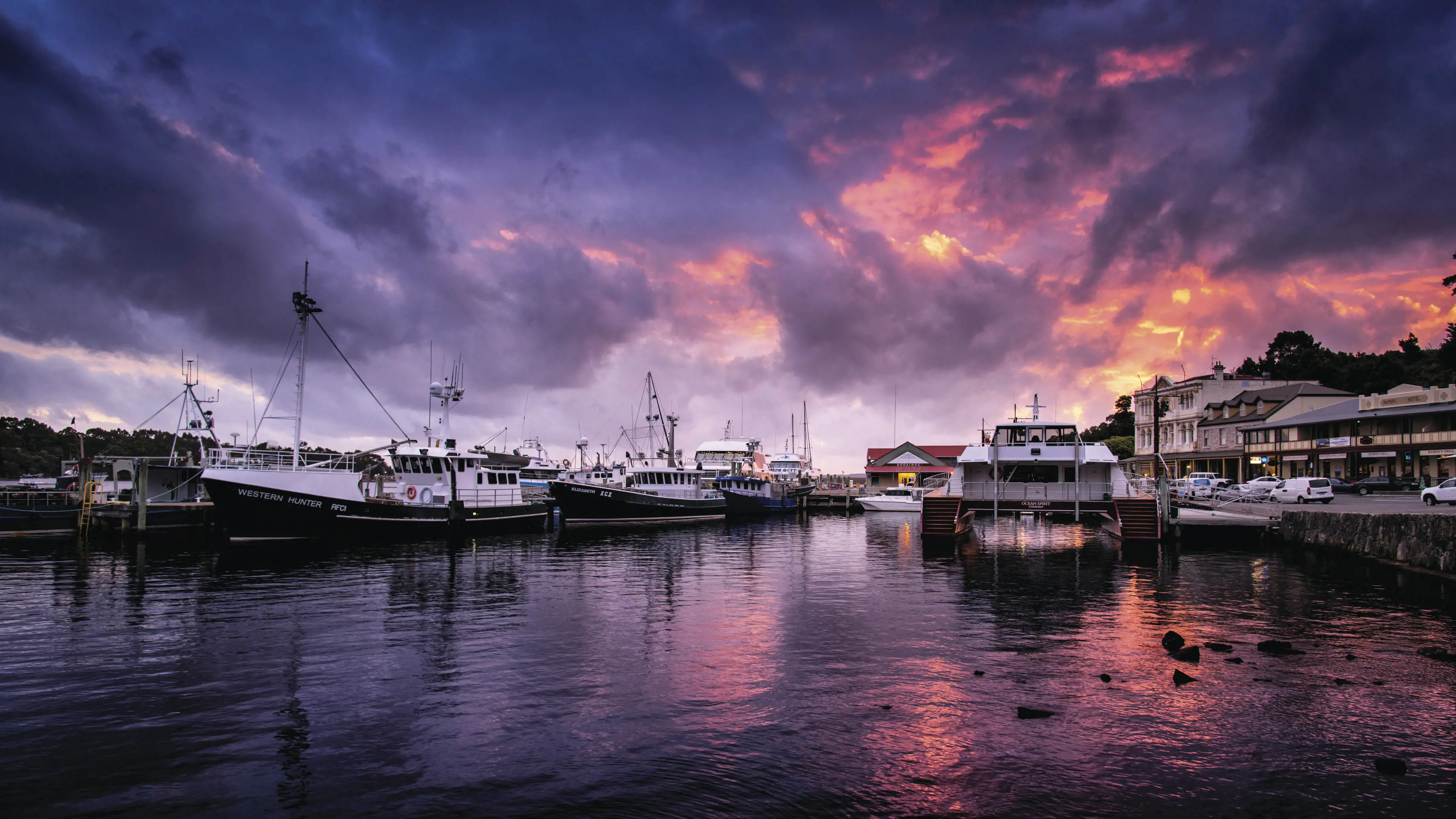 Several boats moored in a harbour. The sky is densely cloudy with rich orange, pink and purple shining through, which is reflected in the water.